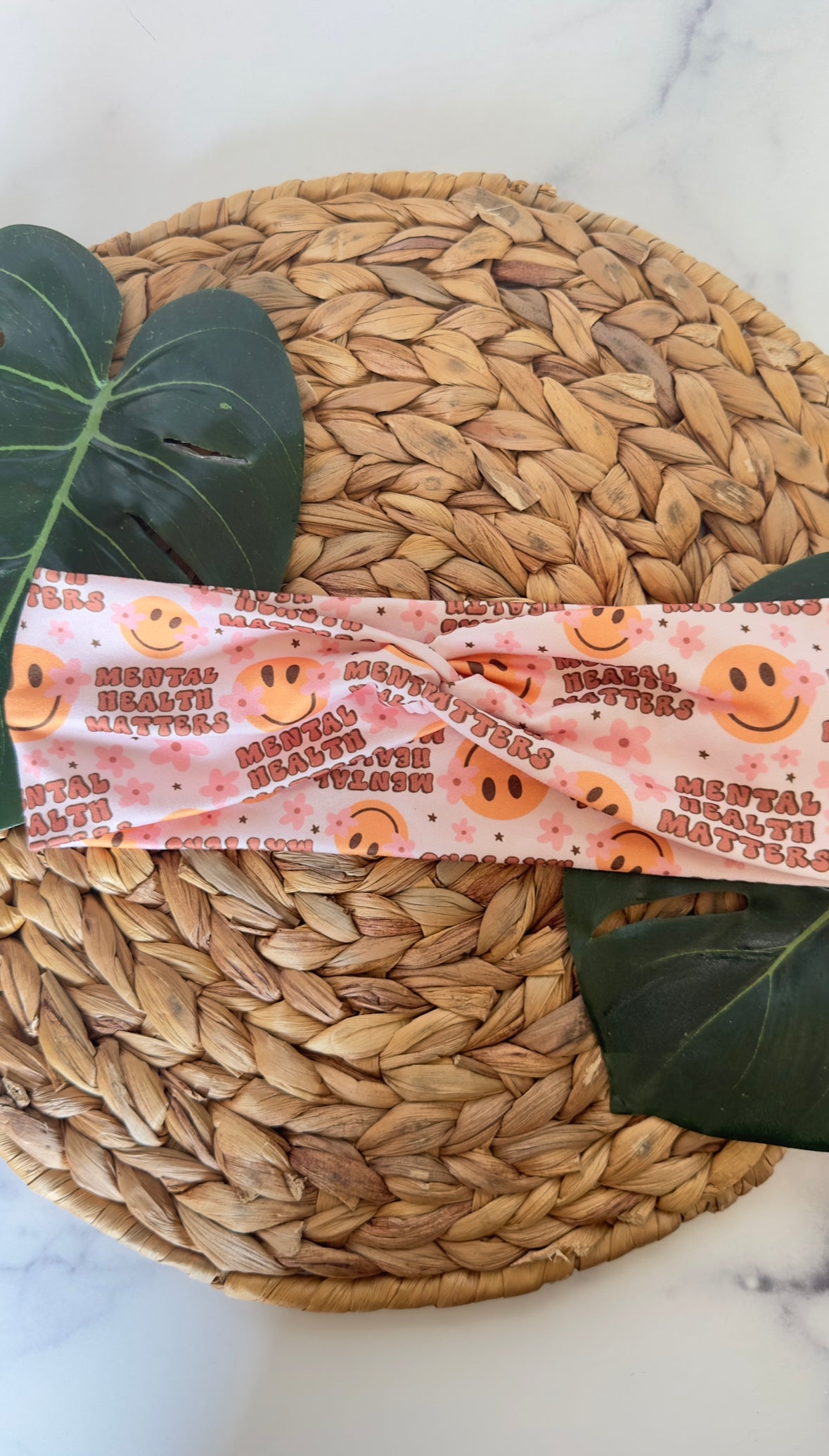 A twist headband on a wicker tray with monstera leaves as background props. The headband features the phrase "mental health matters" in a bubbly wide font. There are smiley faces with pink flowers as dimples.