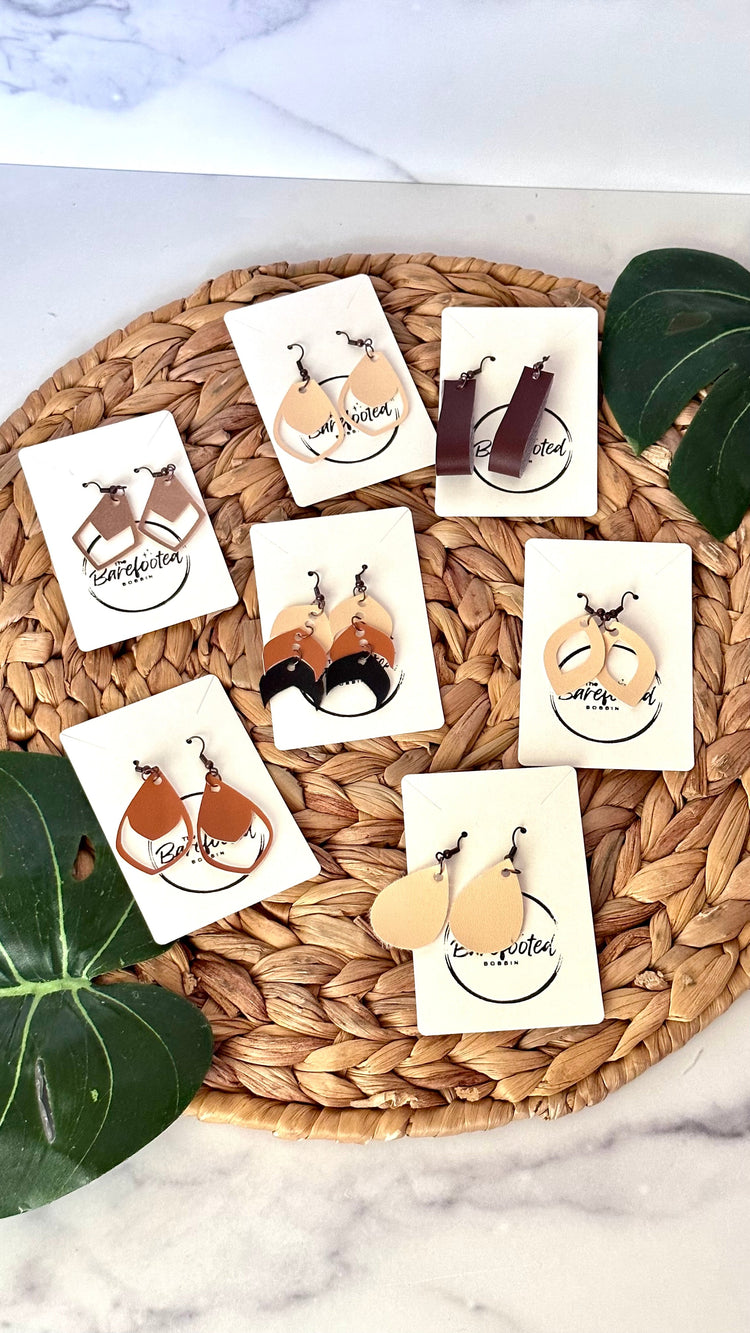 Different colors and styles of faux leather dangly earrings on a round wicker photo background with monstera leaf props