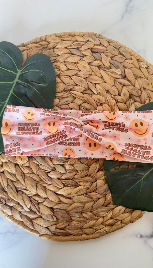 A twist headband on a wicker tray with monstera leaves as background props. The headband features the phrase "mental health matters" in a bubbly wide font. There are smiley faces with pink flowers as dimples. 